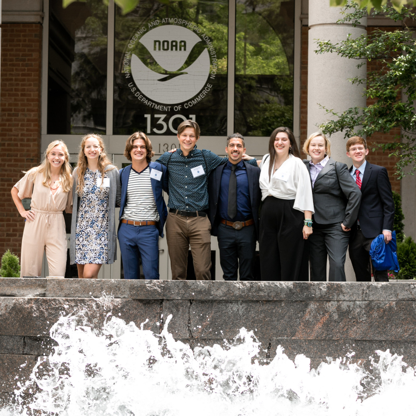 A group of 8 scholars pose outside in a line with their arms around each other. They stand behind a short stone wall that has water splashing against it and in front of a building with the NOAA logo above the door.