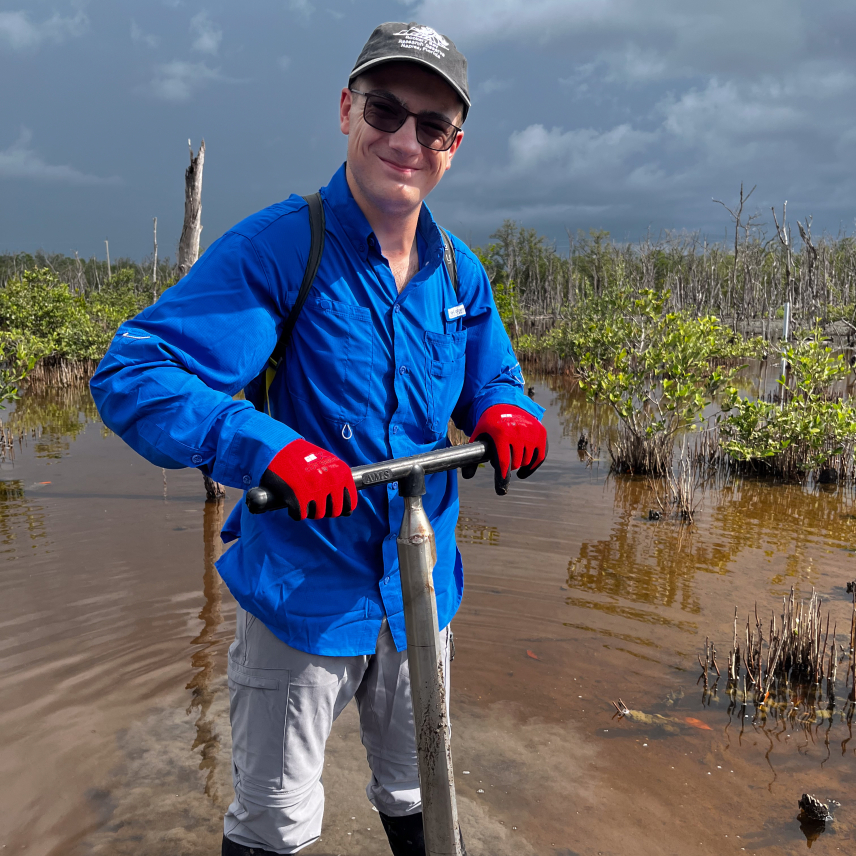 Jonah stands in a wetland with muddied water coming midway up his knee-high boots. He pushes down on a meter-long soil corer and looks at the camera.