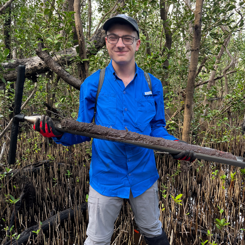 Jonah stands in a mangrove forest, holding a meter-long soil corer that is nearly full with a muddy soil core. 