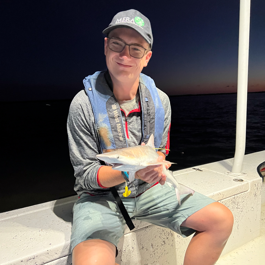 Jonah sits on a boat at night, holding a shark that looks to be about foot and a half long.