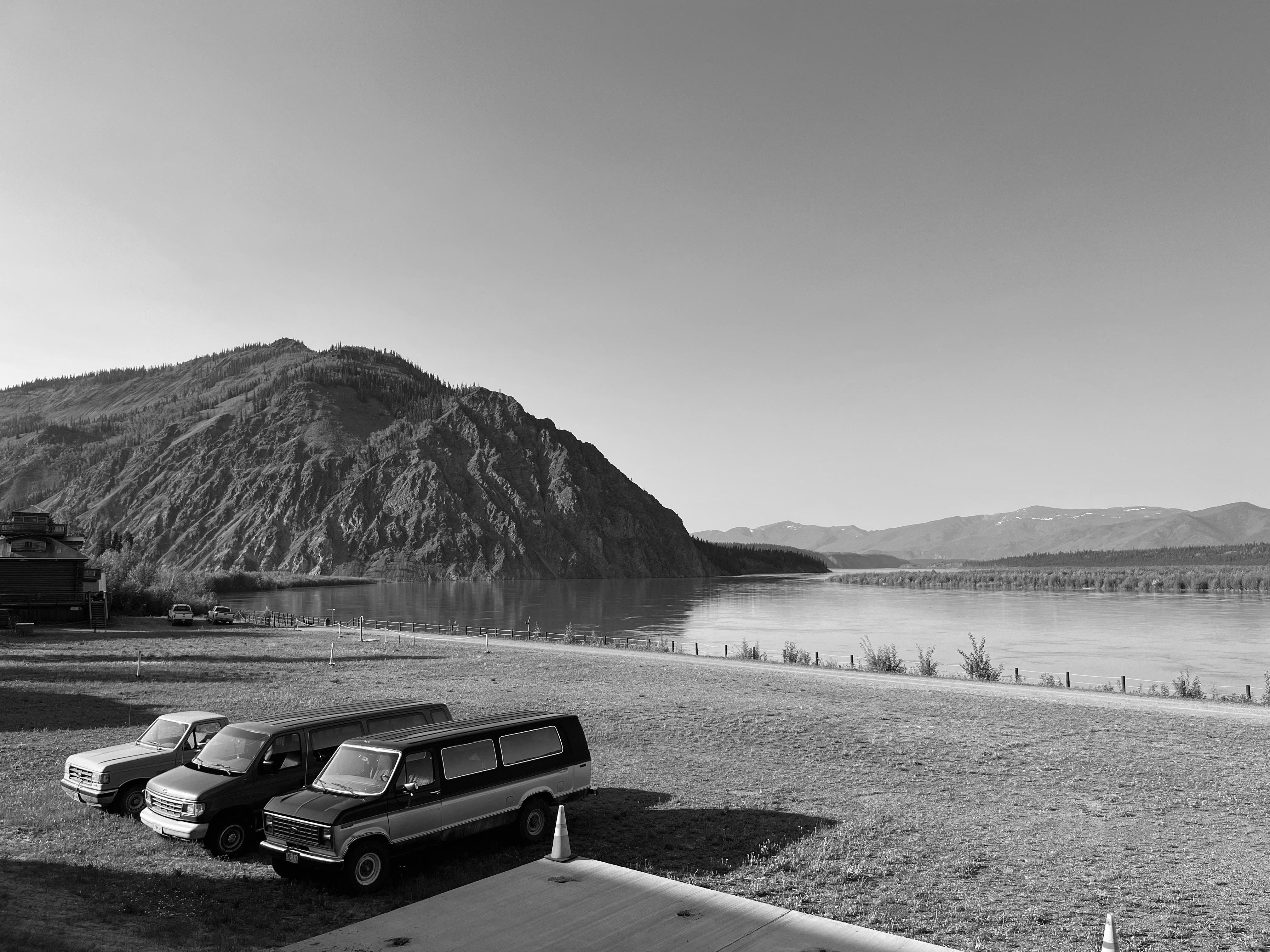 A black and white photo. A nearly empty, grassy parking lot abuts against a wide, calm river. The river bends against a rocky mountain dotted with evergreens and flows into the horizon towards a distant mountain range. A truck and two vans are tucked into the left side of the parking lot, and lend the photo a sense of being taken out of time because they are in good condition, but with a decades old style.