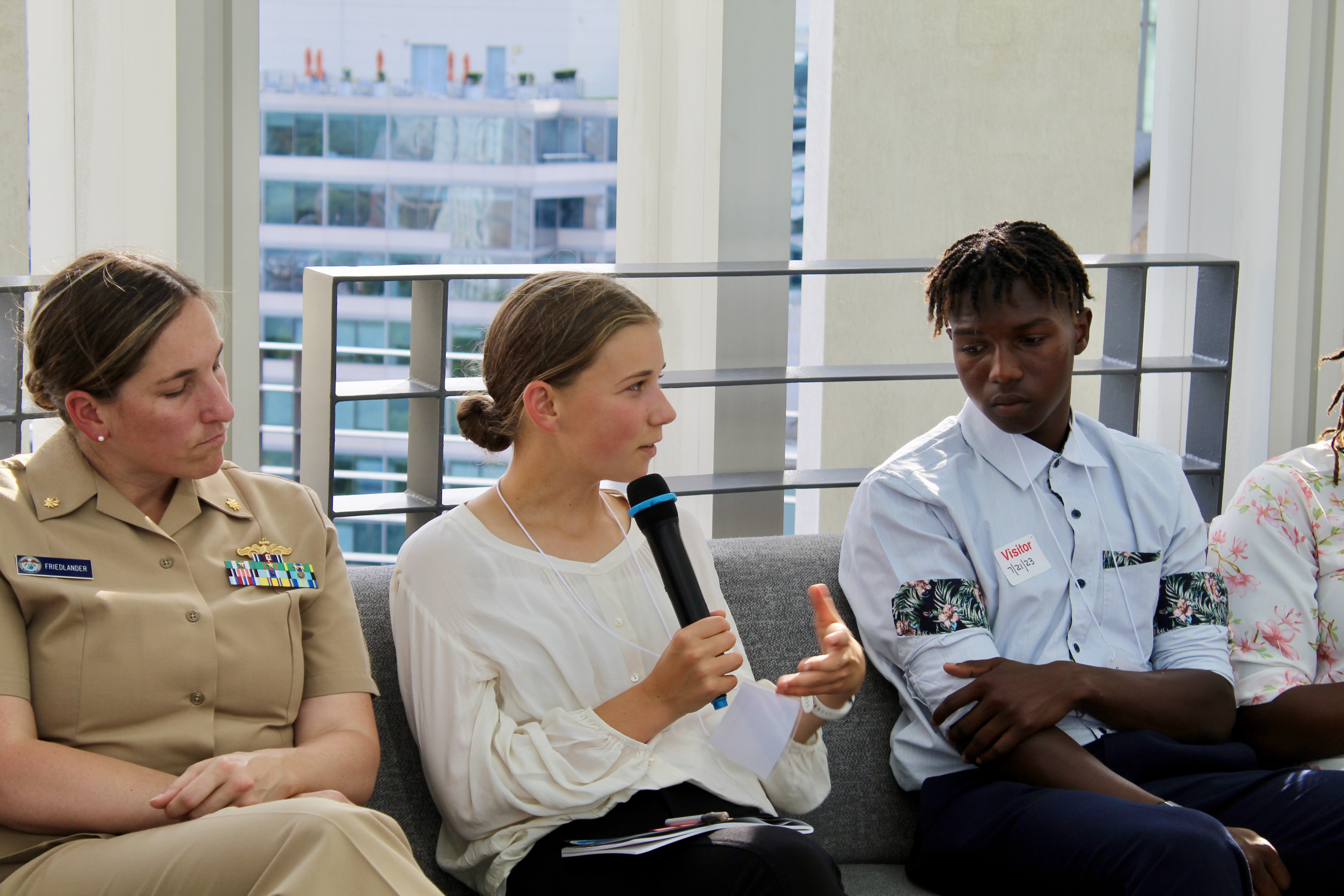 A high school student sits on a couch outside, speaking to the group with a microphone in hand. A NOAA Corps officer and another high school student sit on either side of her.