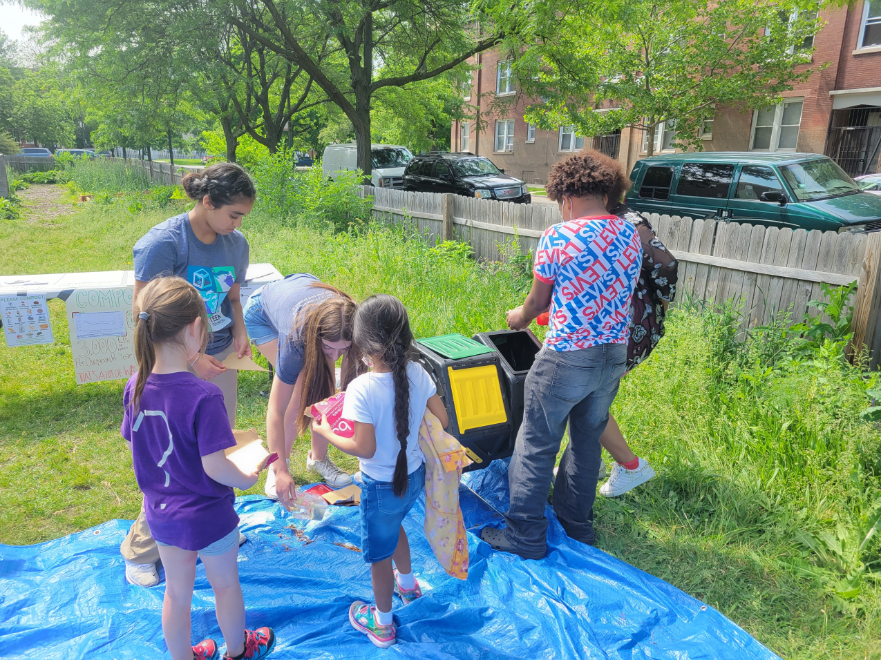 Elementary students watch teens as one gestures toward plastic compost bins and another sorts through small items on a tarp. They are standing in what appears to be a community garden.