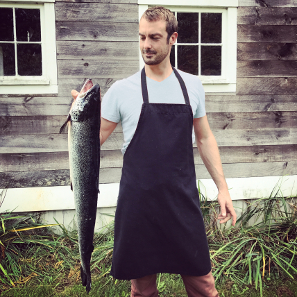 A Chef in a t-shirt and apron holds up a salmon.