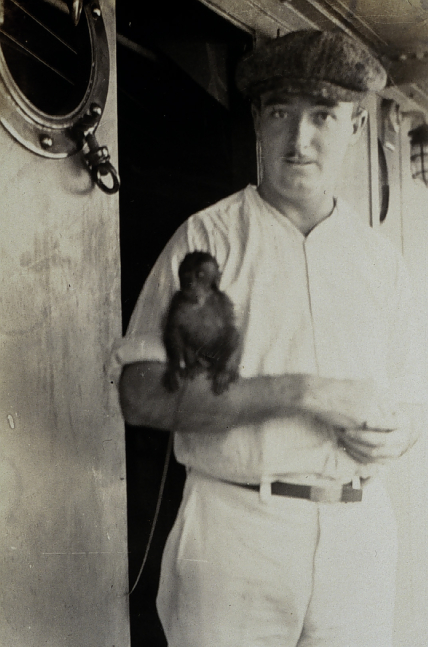 Ken Crosby poses with the USC&GSS Marinduque’s pet monkey. He stands looking forward, with the monkey perched on his arm.