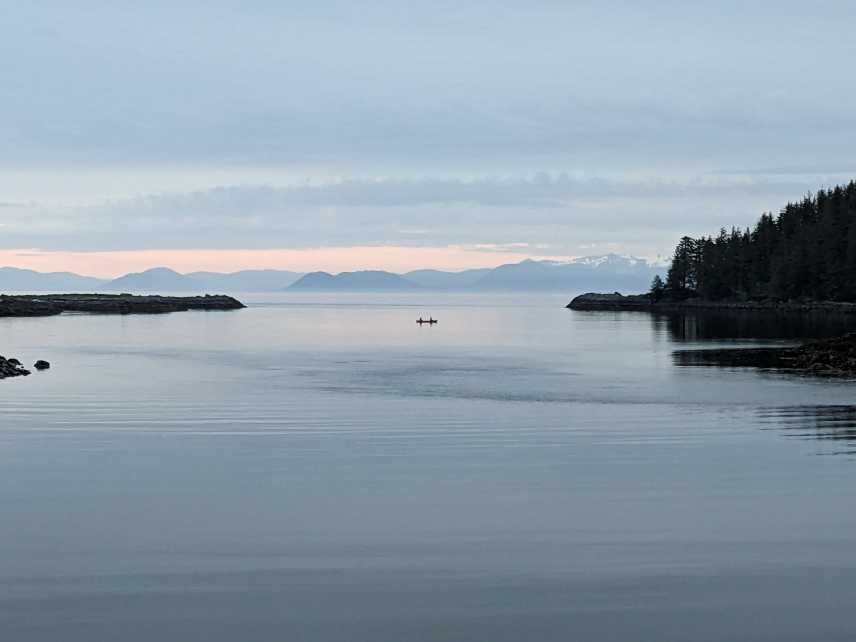 An Alaskan landscape of calm water in an inlet, with a silhouette of two people in a canoe nearly equidistant between land on either side of the inlet. The land on one side is low and flat and the other a rising slop of evergreen trees. In the distance, grey mountainous terrain rests under an overcast sky. The gray and blue tones of the photo create a sense of peace and quiet.