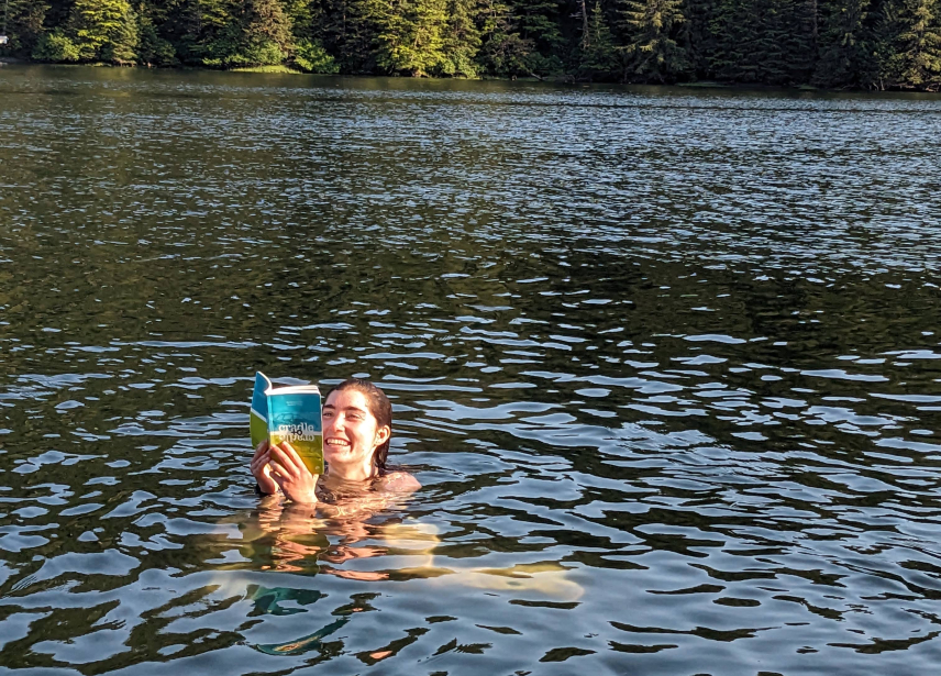 Emma Rudy is outside, submerged up to her neck in a body of water with evergreen trees at the shore across the water. She's smiling and reading a book that she holds just above the water.