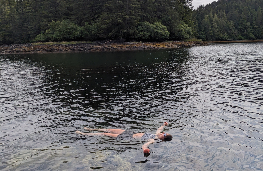 Emma Rudy smiles, floating on her back in shallow water. The nearby island is thick with evergreen trees. She wears swim goggles and her skin looks to be reddened from the cold water.