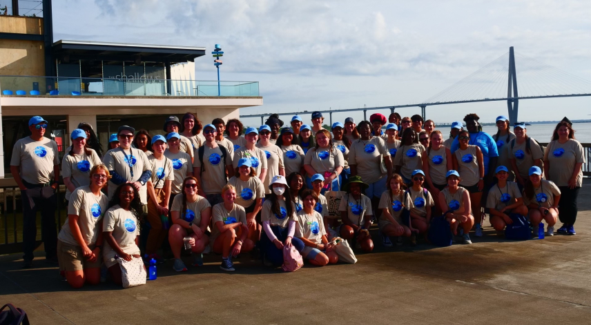 Aquarium teams lined up in 3 rows smiling and wearing matching ‘CELC Youth Summit 2024’ t-shirts and baseball caps with the South Carolina Aquarium deck and a bridge over the water in the background.