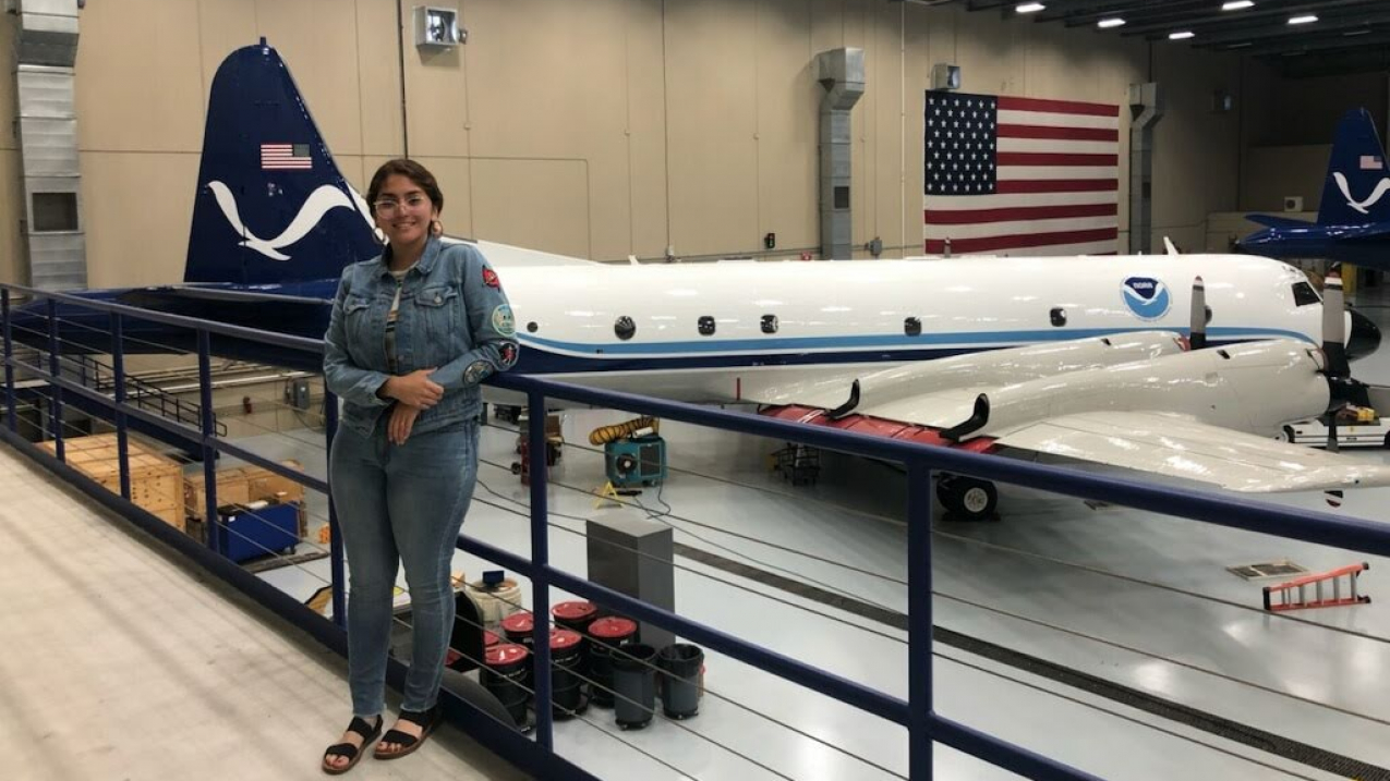 Nohemi poses in a large garage with a plane that has a NOAA Logo