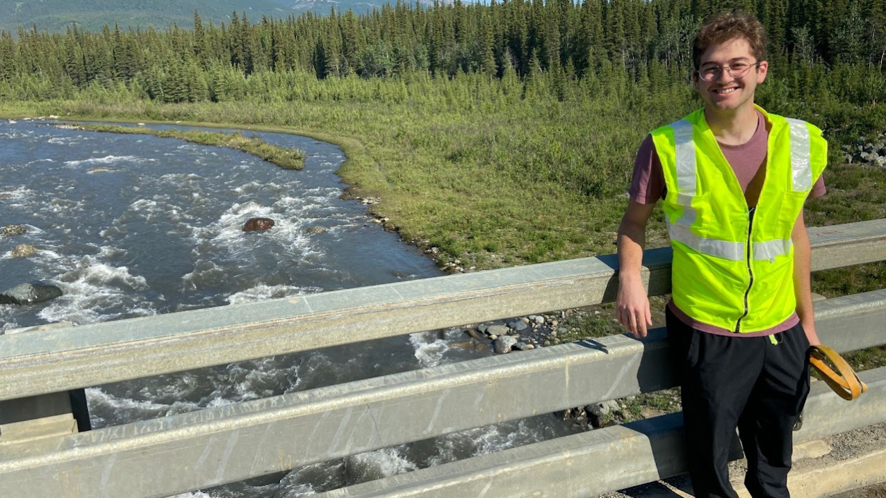 Jacob stands in safety vest on an overpass and smiles at the camera. River rapids flow under the overpass. The river is surrounded by low vegetation that shifts into an evergreen forest, which appears to spread all the way to cover mountains in the distance. 