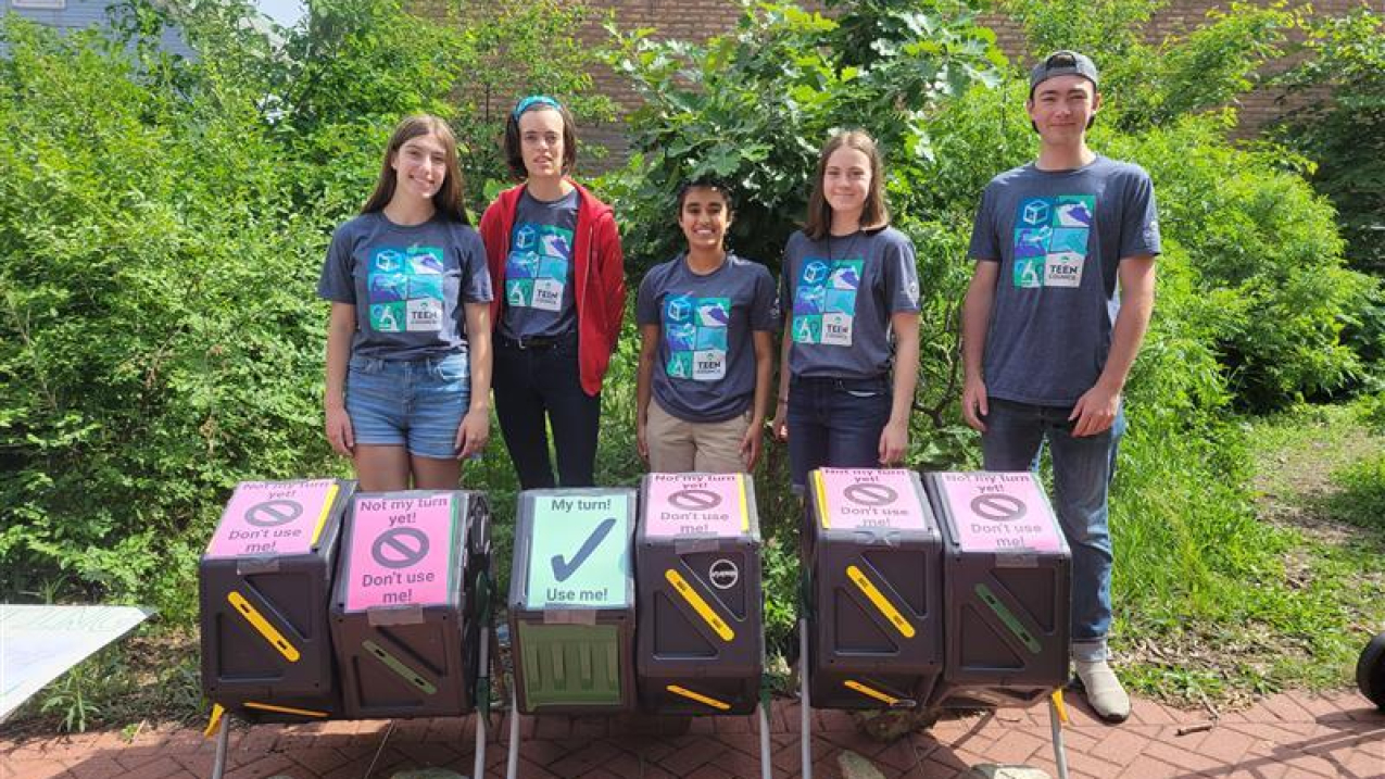 Five teens wearing matching aquarium t-shires stand behind a row of six plastic compost bins. Five of the bins have signs on top that say, "Not my turn yet! Don't use me!" while one says, "My turn! Use me!" They are outdoors, surrounded by lush greenery.