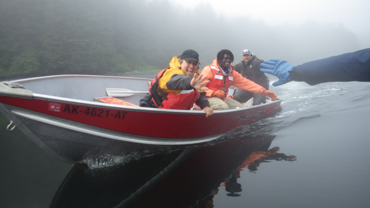 Three people in a row boat grin and lean over the side, each stretching an arm out towards a hand that can be seen coming into the side of the photo. The day looks to be calm and foggy and everyone wears personal floatation devices over their cold-weather field clothes.