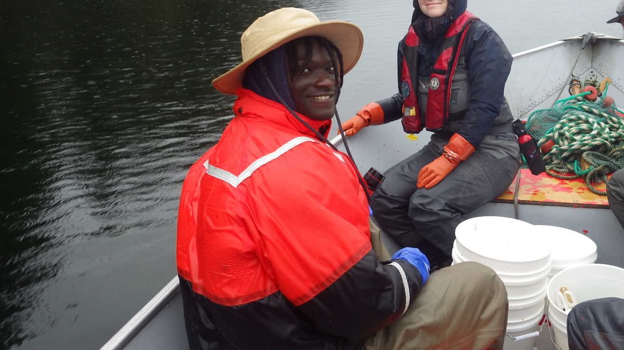 TreVaughn smiles, sitting on an aluminum fishing boat loaded with field equipment. He's dressed for aquatic fieldwork in cold weather. 