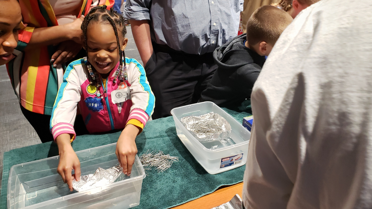 A child looks excitedly at their aluminum foil boat in a tub of water.