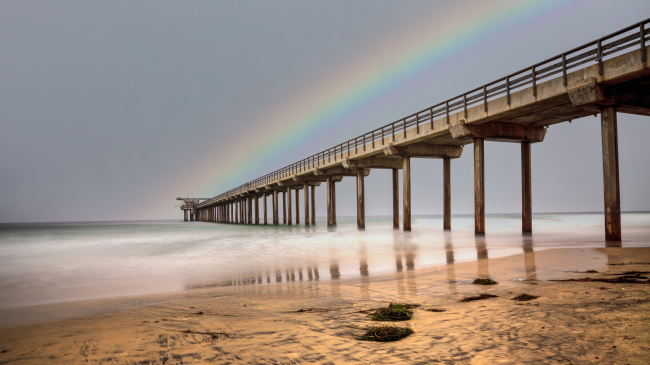 A rainbow over a partly cloudy Scripps Beach and pier in La Jolla, California, during Summer 2020