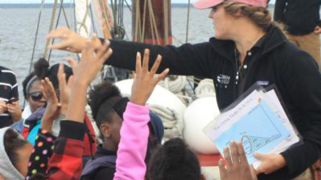 Students are on a boat in a river raising their hands as an educator talks to them.