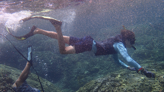 Samuel swims in snorkeling gear near to the water's surface against a background of rock and corals. The leg of a second snorkeler can be seen as the person swims out of the shot. 