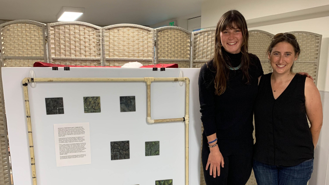 Kate and Nicole stand together with one arm around each other. They are smiling and standing next to a display of paintings, which are nested inside of a 1 meter by 1 meter square made of pvc (a sampling quadrat).