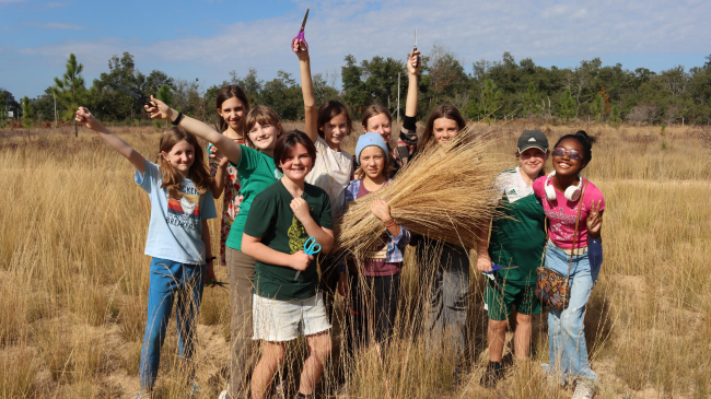 Ten children stand together in a field of golden wheat-like wiregrass on a bright sunny day. Several of the students are holding scissors above their heads victoriously, while one child stands in the middle of the group holding a large armful of wiregrass stalks in one arm. All the students have smiles of success on their faces, and one child on the edge of the group wearing sunglasses flashes the peace sign.