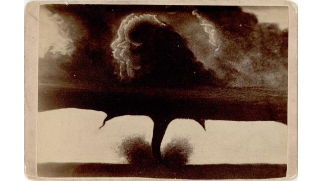 A black and white photo of a tornado with multiple vortices, one in the center, which reaches to the ground and a smaller one on either side which do not reach the ground.