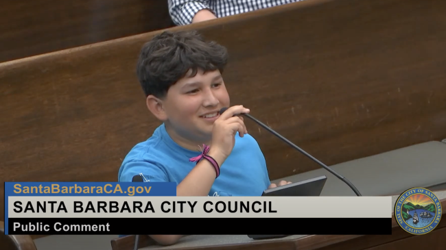 A young boy in a blue Ocean Guardian T-shirt stands at a lectern with a microphone speaking to the mayor and City Council of the City of Santa Barbara. 