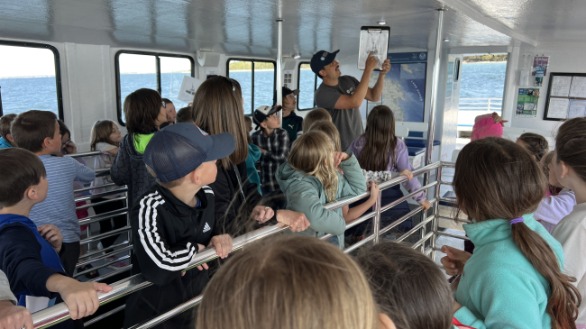A third grade class looks at an educator holding a photo while aboard a boat.