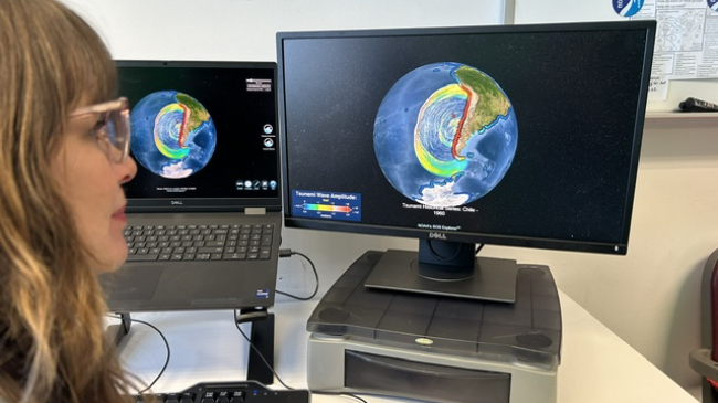 A person sits at a desk looking at a laptop screen and a computer monitor. The screens show a globe displaying a tsunami dataset in SOS Explorer.