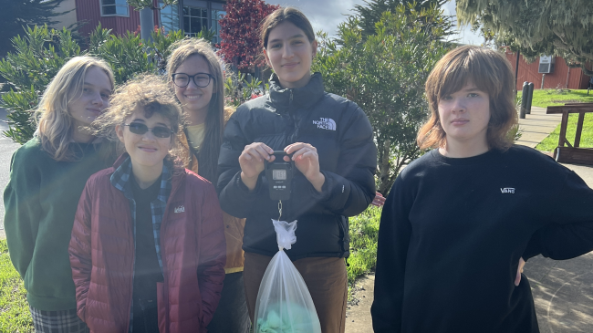  Five students from Chartwell School stand behind their yellow Ocean Guardian Go Bags (backpacks). The student in the center of the image weighs a bag full of collected trash. 