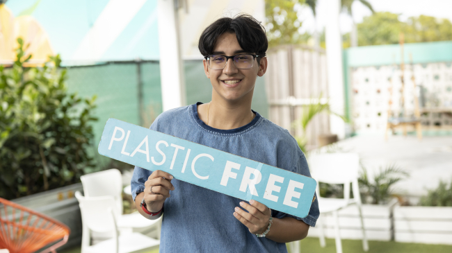 A teenaged Junior Sustainability Consultant smiles while holding a sign that reads "Plastic Free" in an outdoor patio area. 