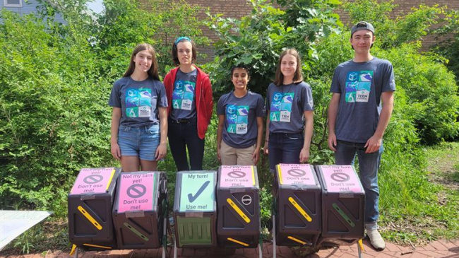 Five teens wearing matching aquarium t-shirts stand behind a row of six plastic compost bins. Five of the bins have signs on top that say, "Not my turn yet! Don't use me!" while one says, "My turn! Use me!" They are outdoors, surrounded by lush greenery.