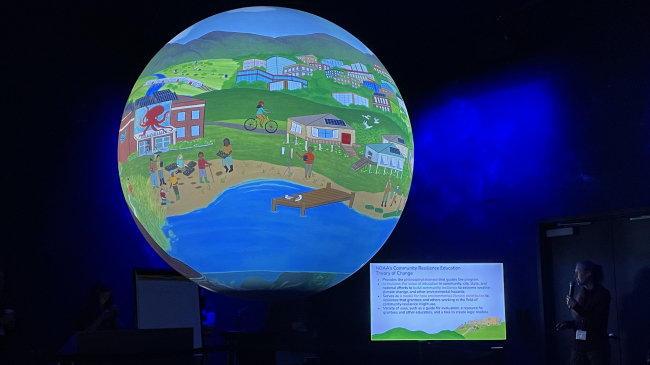 A large sphere with a cartoon coastal community projected onto it and a speaker to the side, describing the images.