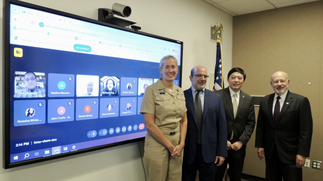 VADM Hann, Ben Friedman, Dr. Sen Chaio, and Dr. Rick Spinrad pose for a photo standing next to a virtual meeting that includes other members of NOAA and the EPP/MSI Cooperative Science Centers. The U.S. flag is behind them. 