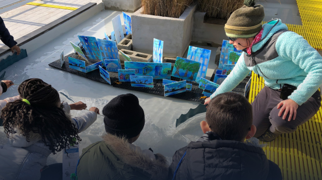 Three students use pieces of plastic and board to push water towards a replica of Norfolk. An instructor assists the students with the activity.