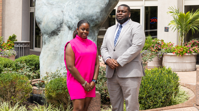 Two people in business formal clothing pose for a photo outside in front of a large statue of a hand.