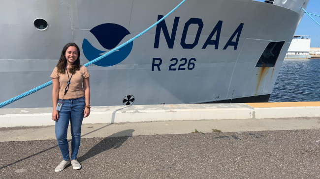 Paola stands in front of a large white vessel. The ships side displays the NOAA logo and "NOAA" signage. 