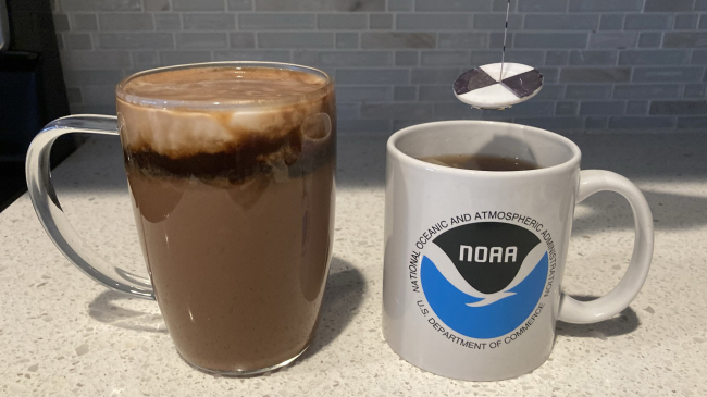 Two mugs are placed on a kitchen counter. One mug has the NOAA logo on it and a miniature secchi disk is suspended above it. (The secchi disk is made of a white disk with black on opposite quarters. It is hanging from a string that has markings every centimeter.) The other mug is made of clear glass, and it contains hot cocoa topped with a layer of espresso and a layer of milk. The top two layers are mixing, forming swirls.