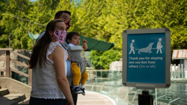A masked family at an aquarium looks at something off-camera, standing in back of a sign that reads, “Thank you for staying one sea lion from others (2 m or 6 ft).”
