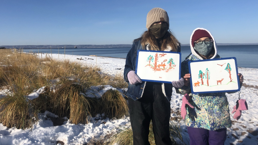 Two students stand on a snow-covered sand dune on the edge of a lake holding whiteboards with drawings that appear to include conifer trees, foxes, deer, and eagles. The students are dressed for winter weather and are both wearing face masks.