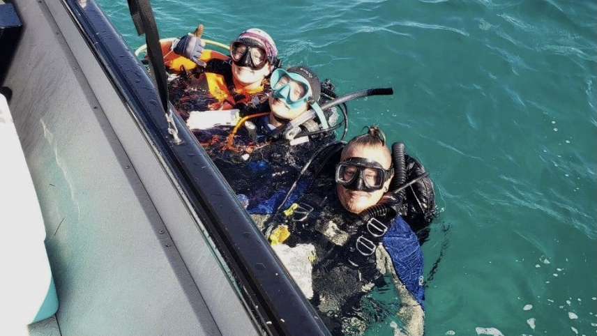 Three people in full dive gear float in the water and hang onto the side of a boat, posing for the camera.