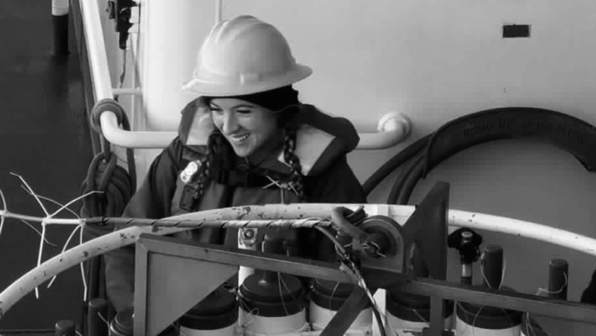 A black and white photo of Devynn aboard a research vessel. She wears a hardhat along with cold weather field clothes and appears to be interacting with large circular piece of equipment with many cylinders attached around its circumference.