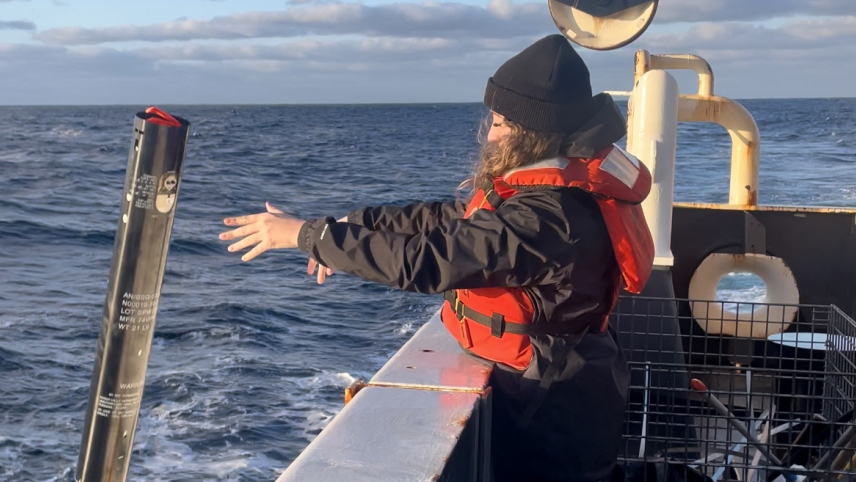 Devynn is caught mid-action, with arms outstretched as she tosses a sono-bouy over the side of a research vessel. She wears cold weather field clothes and a life jacket.