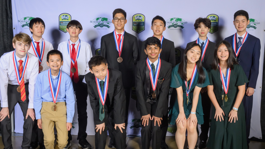 Twelve students dressed in formal attire pose for a group photo in front of a backdrop that has the 40th Science Olympiad National Tournament logo on it.