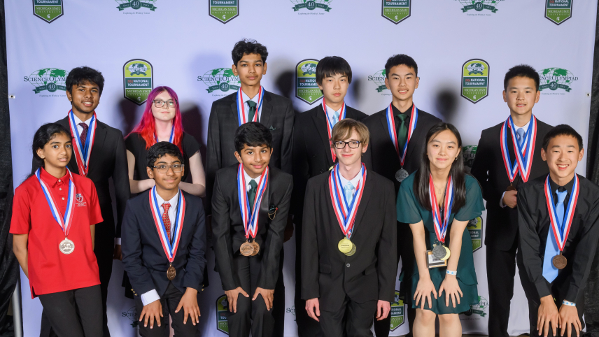 Twelve students dressed in formal attire pose for a group photo in front of a backdrop that has the 40th Science Olympiad National Tournament logo on it. 