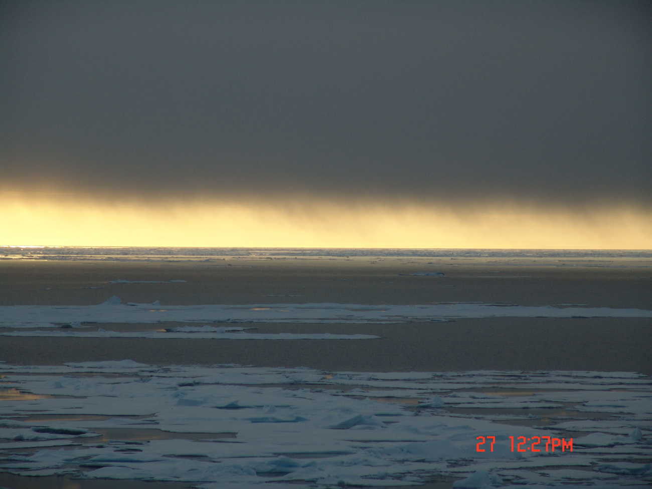 A dark blanket of cloud framing a yellow-gold sun illuminated horizon withice floes and a polynya below