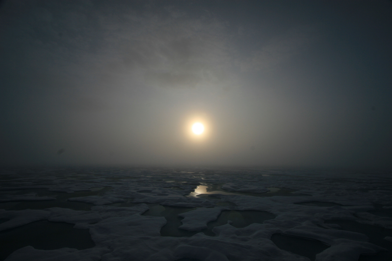 Sun seen through the clouds over melt pools and hummocky ice on a gloomyday
