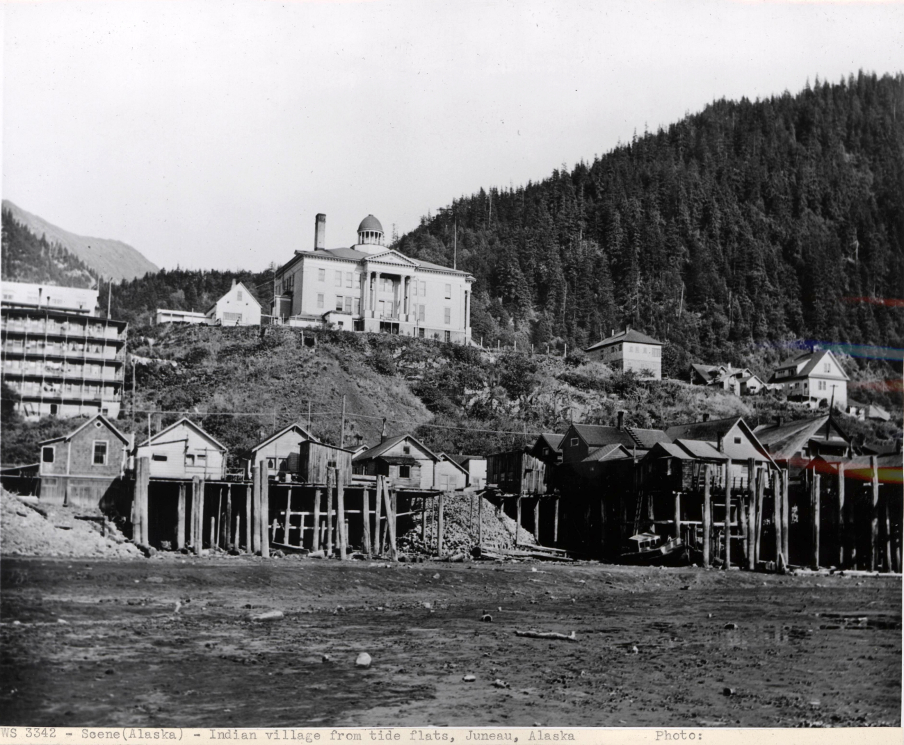Indian village on stilts on tidal flats with United States Courthouse on hillabove