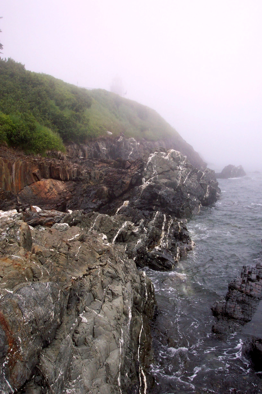 Quartz-veined metamorphic rocks meet the sea below a fog-shroudedlighthouse at West Quoddy Head
