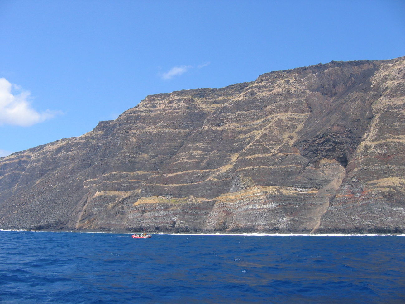 A Safeboat from the NOAA Ship OSCAR SETTE is dwarfed by multiplelava flows forming a cliff on the coast of Hawaii