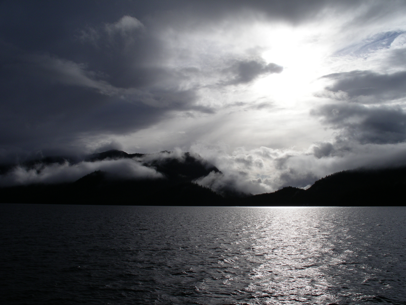 A dramatic view of clouds, fog, coastal mountains,and a cloud obscured sun shining off a grey ocean